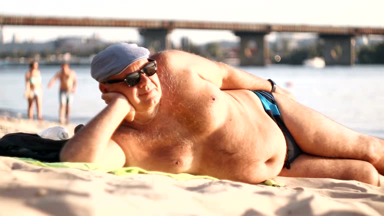 Senior man resting, lying on sand at the beach.  Summertime travel vacation at the beach. Senior man body on beach In summer. Man in swimming trunks and a cap sunbathing on the beach.